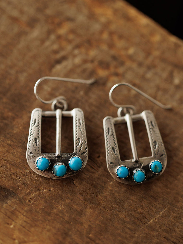 The Los Alamos Earrings by Savannah Sevens western life{&}style feature sterling silver horseshoe buckles with three round turquoise stones at the base, shown on a rustic wooden surface.