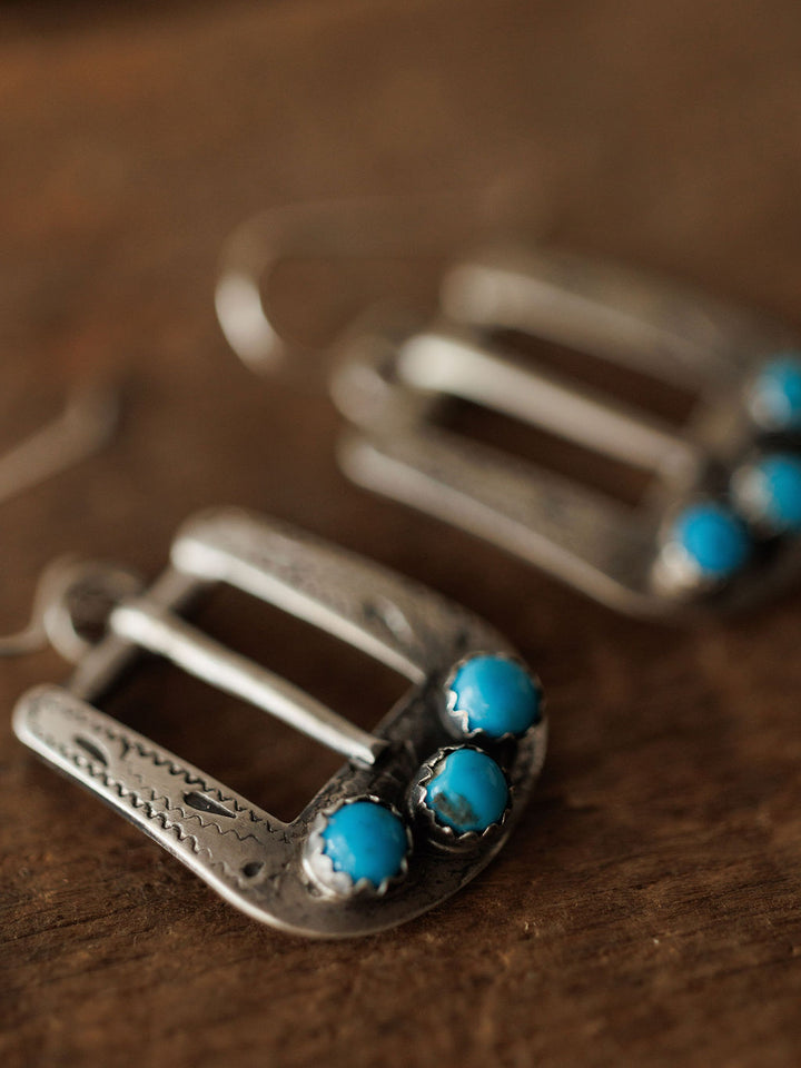Close-up of the Los Alamos Earrings by Savannah Sevens western life{&}style, sterling silver and shaped like horseshoe buckles, each set with two round turquoise stones at the bottom and displayed on a wooden surface. Another earring is softly blurred in the background.