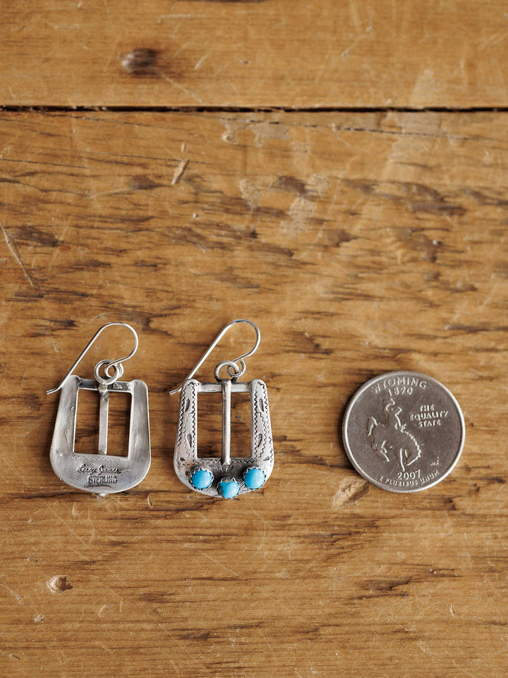 The Los Alamos Earrings by Savannah Sevens western life{&}style, sterling silver with blue stones, are shown on a wooden surface beside a Wyoming state quarter for size. One earring displays the engraved back and the other the front.