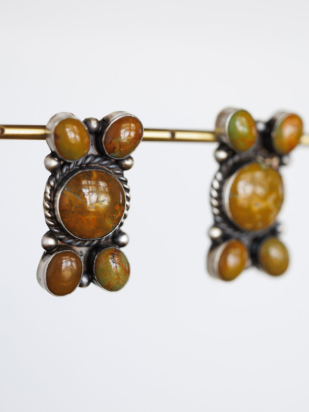 The Yuma Earrings by Savannah Sevens western life{&}style feature vintage-inspired handcrafted sterling silver with round and oval amber stones in ornate frames, displayed hanging on a brass rod against a white background.
