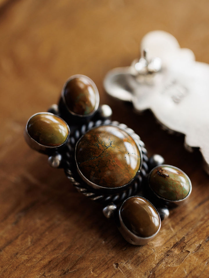 The Yuma Earrings by Savannah Sevens western life{&}style feature handcrafted sterling silver with five polished brown and green stones in ornate metalwork, beautifully displayed on a wooden surface.