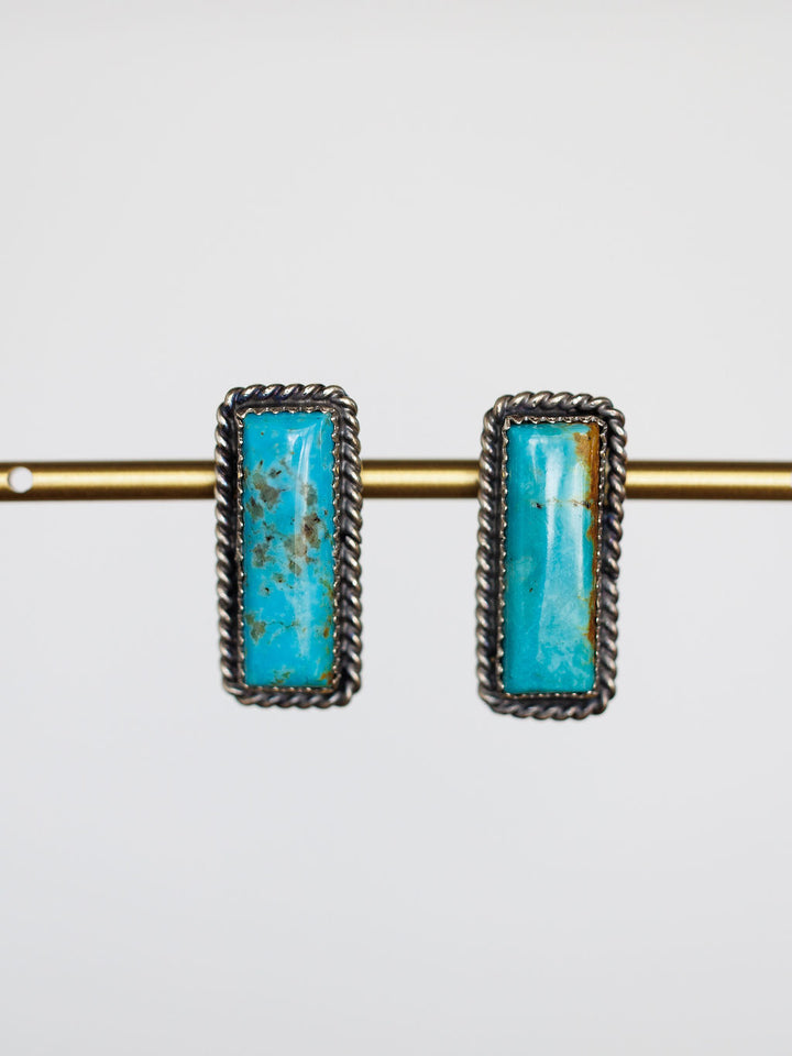Toano Earrings by Savannah Sevens western life{&}style: Navajo handcrafted turquoise bar earrings with twisted sterling silver borders, shown hanging on a brass rod against a white background.