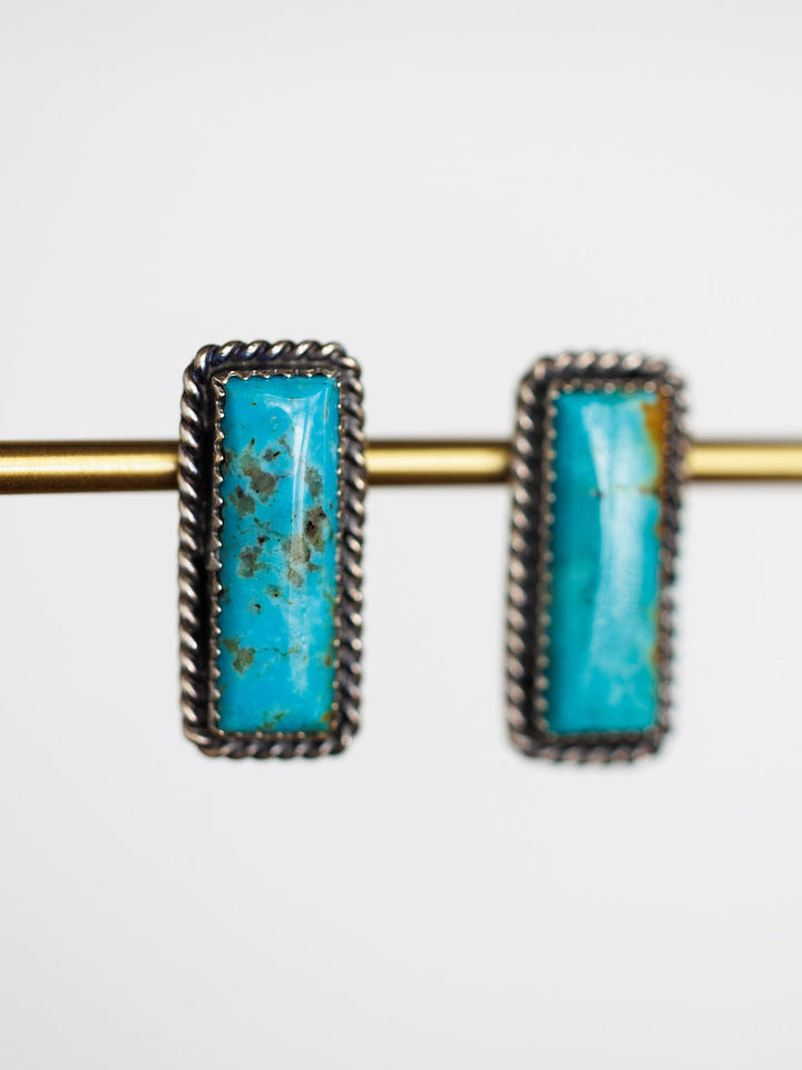 The Toano Earrings by Savannah Sevens western life{&}style are Navajo handcrafted turquoise bar earrings with sterling silver twisted rope borders, shown hanging from a gold rod on a white background.
