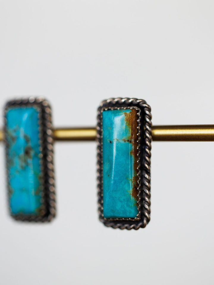 Close-up of Toano Earrings by Savannah Sevens western life{&}style: turquoise bars with twisted silver borders and sterling details, hanging on a brass rod against a plain white background. Navajo handcrafted with elegant rectangular stones.