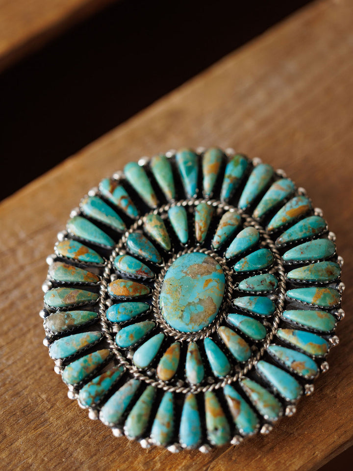 The Holyoke Buckle by Savannah Sevens western life{&}style is a round, silver piece featuring concentric rows of Kingman turquoise teardrop and oval stones, shown on a wooden surface.