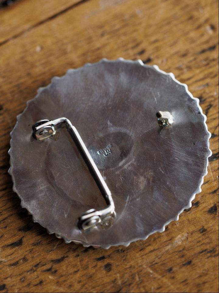 A close-up of the back of the Holyoke Buckle from Savannah Sevens western life{&}style, featuring a round sterling silver design with a bar and hook attachment, lightly textured edges, and subtle center markings, resting on wood.