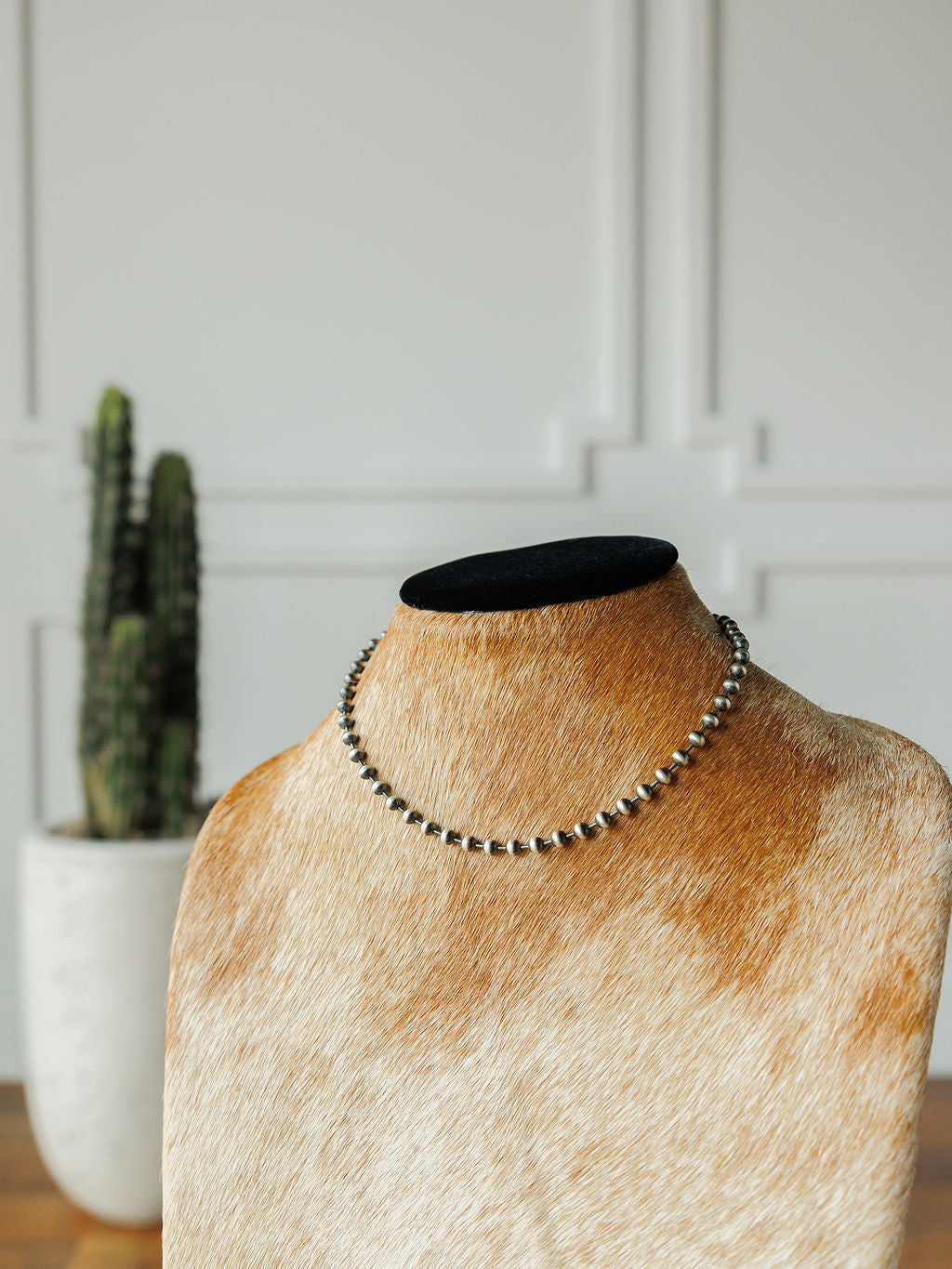 The Sedalia Necklace by Savannah Sevens western life{&}style is showcased on a cowhide-covered bust with a black velvet top, while a potted cactus sits in the background against a white paneled wall.