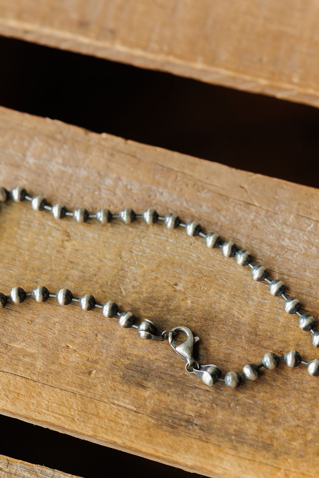 A close-up of the Sedalia Necklace by Savannah Sevens western life{&}style, a sterling silver beaded chain with a lobster clasp inspired by Navajo pearl designs, shown on a textured wooden surface.