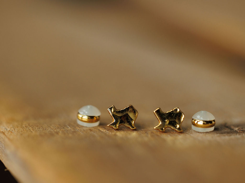 Two pairs of small stud earrings from Savannah Sevens western life{&}style: the Texas Earrings in gold star shapes and a pair with round white stones set in gold, both displayed on a wooden surface with a softly blurred background.