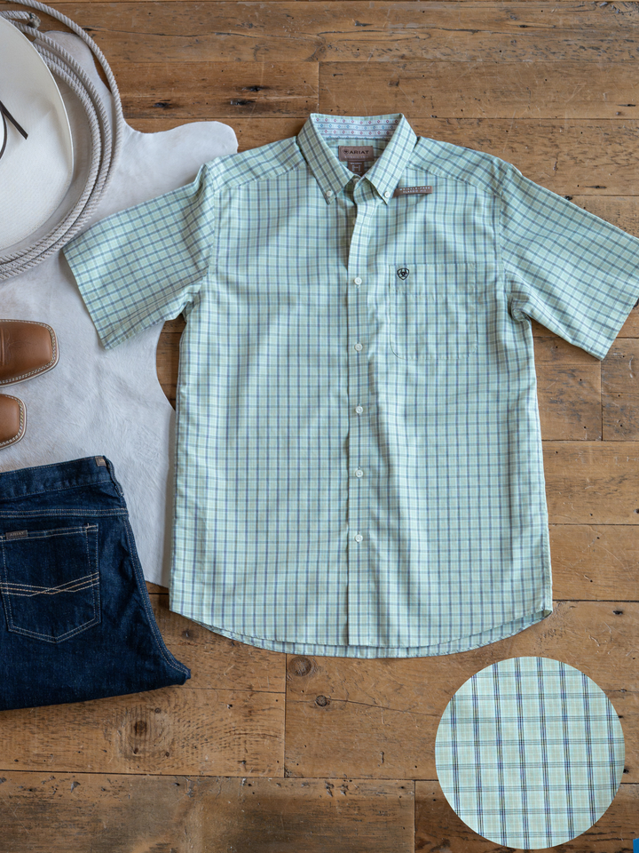 The Graham {Men's} by Savannah Sevens western life{&}style, a light green and blue plaid short sleeve button-up shirt, is displayed with jeans, a white hat, belt, and brown shoes. A circle highlights its wrinkle-free fabric pattern.