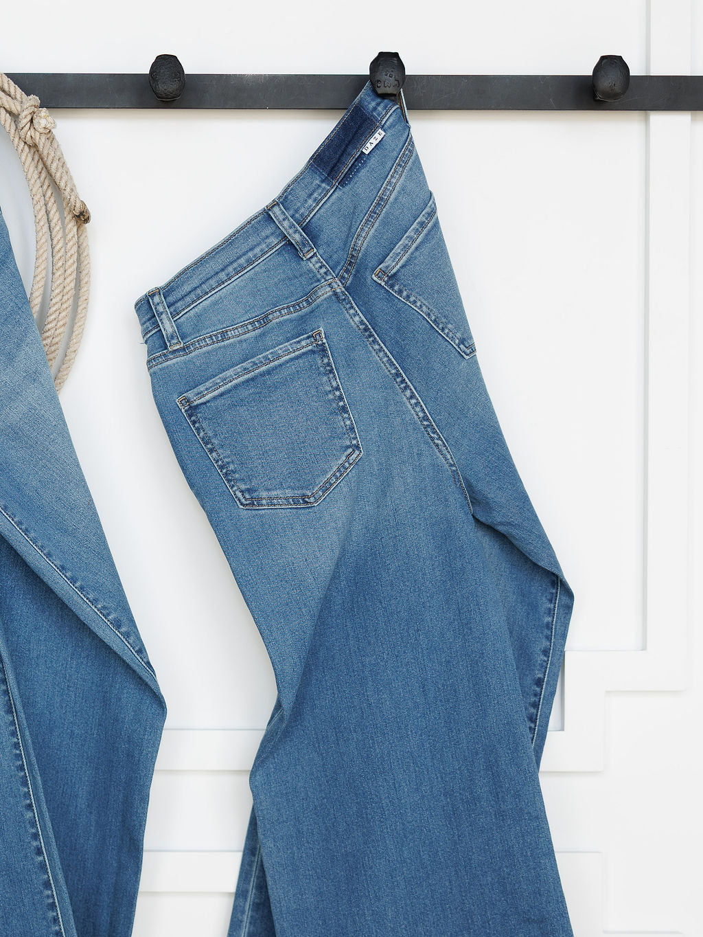 The Jacey High Rise Wide Leg Jean by Savannah Sevens western life{&}style in light blue hangs on a black hook against a white wall, with part of another denim piece and a rope visible to the left.