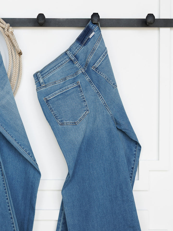 The Jacey High Rise Wide Leg Jean by Savannah Sevens western life{&}style in light blue hangs on a black hook against a white wall, with part of another denim piece and a rope visible to the left.