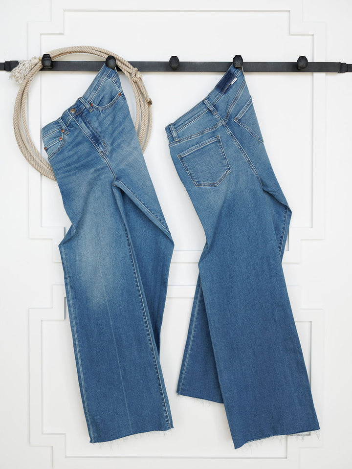 Two pairs of Savannah Sevens western life{&}style’s The Jacey High Rise Wide Leg Jeans in medium wash hang side by side on a black rod against a white wall, with a thick rope looped around the left end of the rod.