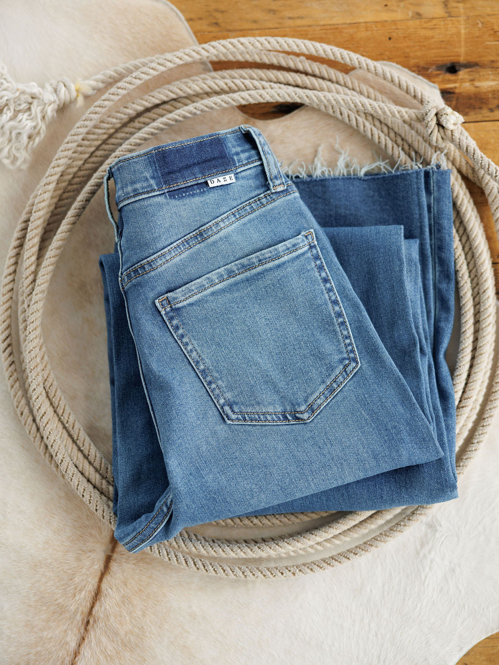 A pair of Savannah Sevens western life{&}style The Jacey High Rise Wide Leg Jeans is neatly folded inside a coiled rope, resting on a light cowhide rug atop a wooden floor.