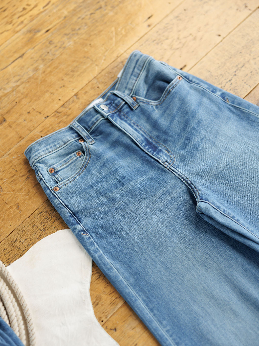 A pair of The Jacey High Rise Wide Leg Jeans by Savannah Sevens western life{&}style in light blue lies flat on a wooden floor beside a white faux fur rug and a coiled rope.
