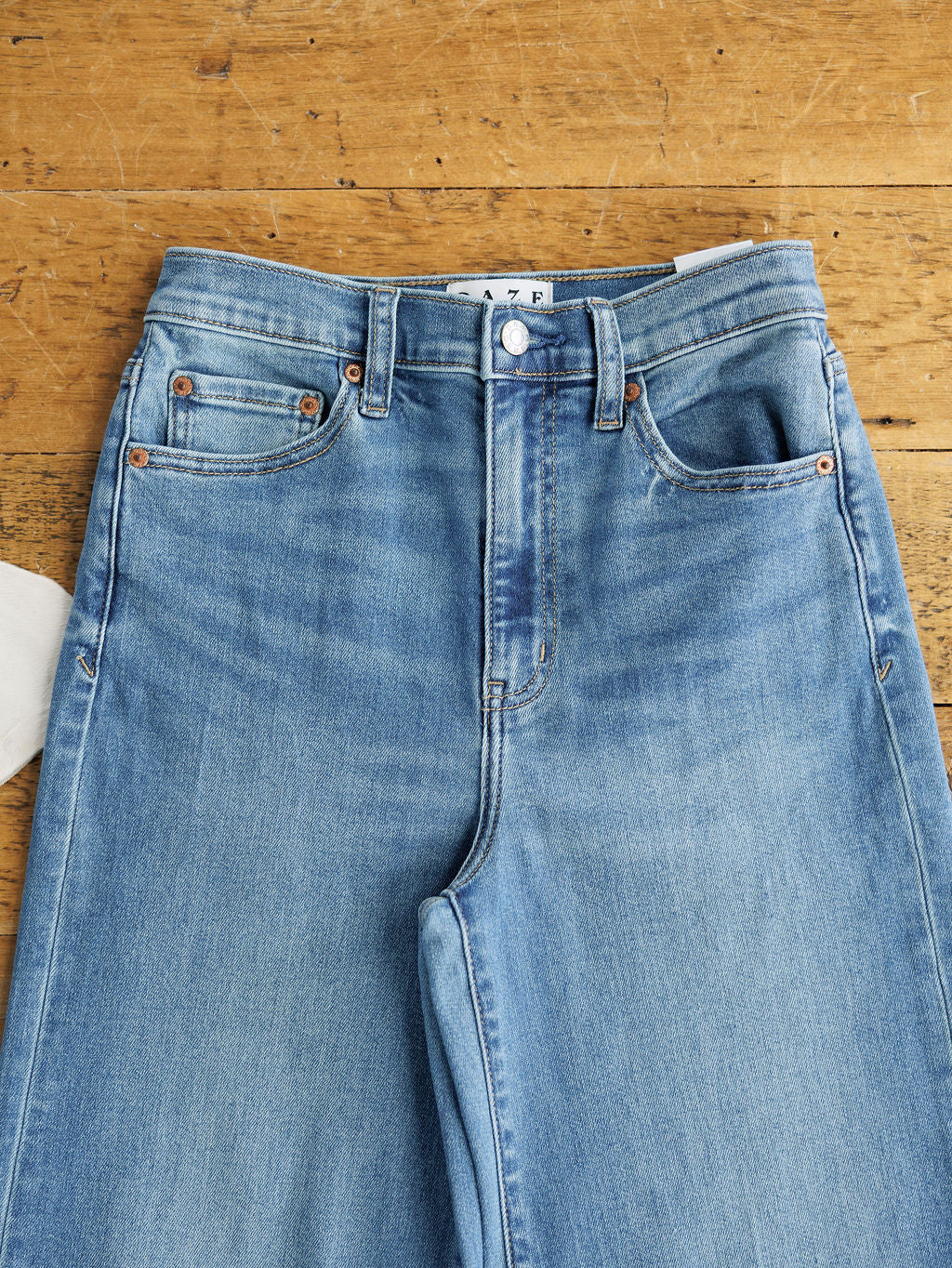 The Jacey High Rise Wide Leg Jean by Savannah Sevens western life{&}style displayed flat on a wooden surface, showcasing the medium wash fabric, waistband, front pockets, and wide leg design.