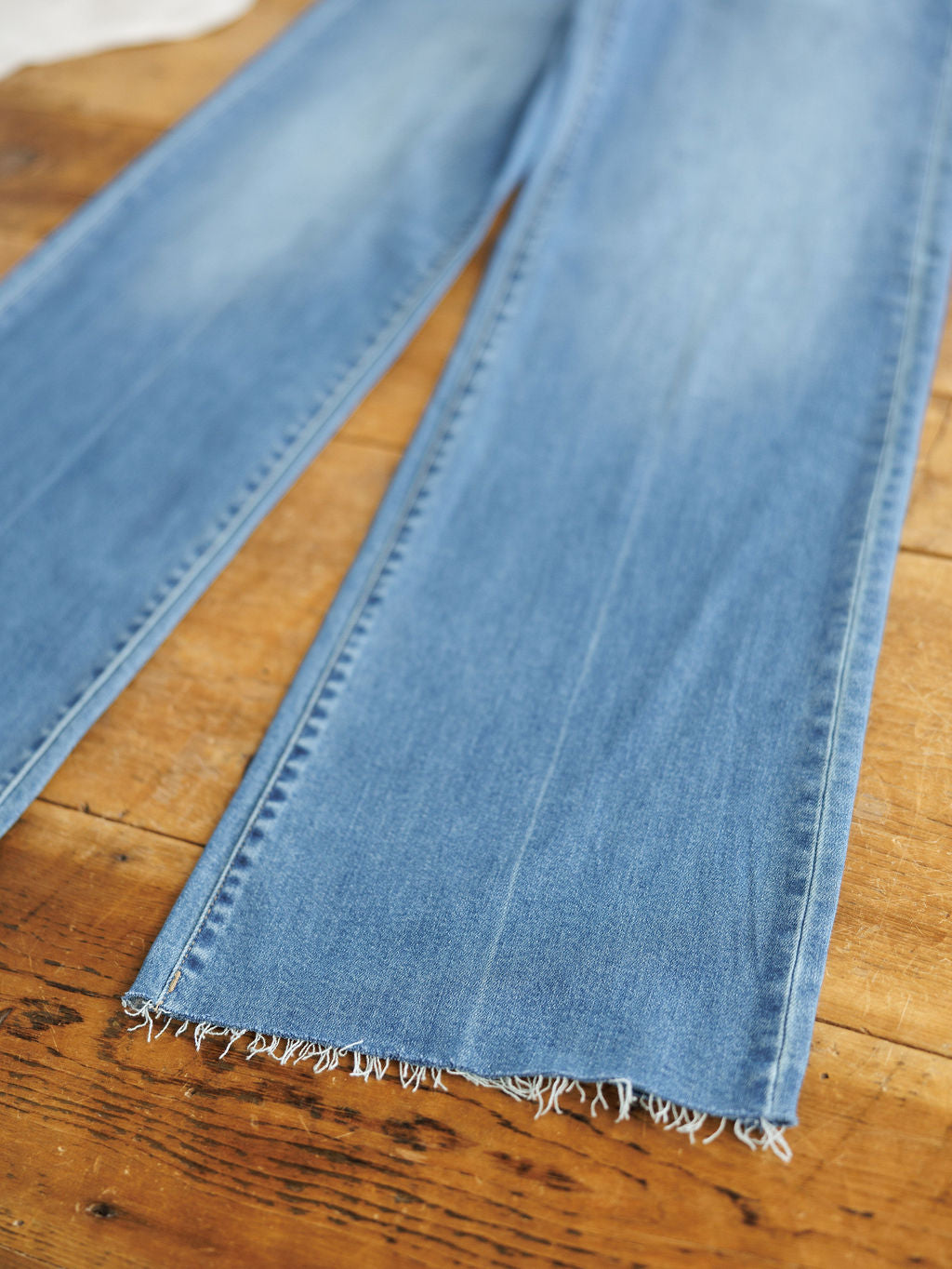 The Jacey High Rise Wide Leg Jean by Savannah Sevens western life{&}style in light blue, featuring frayed, unfinished hems, is shown laid flat on a wooden surface.