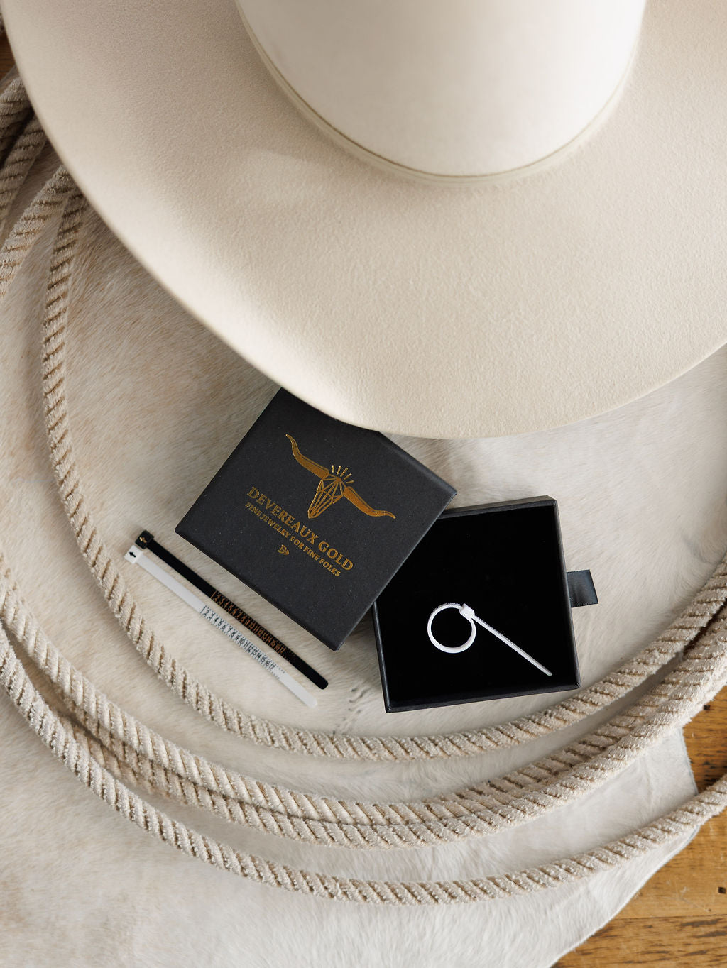 A beige cowboy hat, white pen, and black jewelry box with a silver ring are set on a light cowhide rug beside a Devereaux Gold branded box.