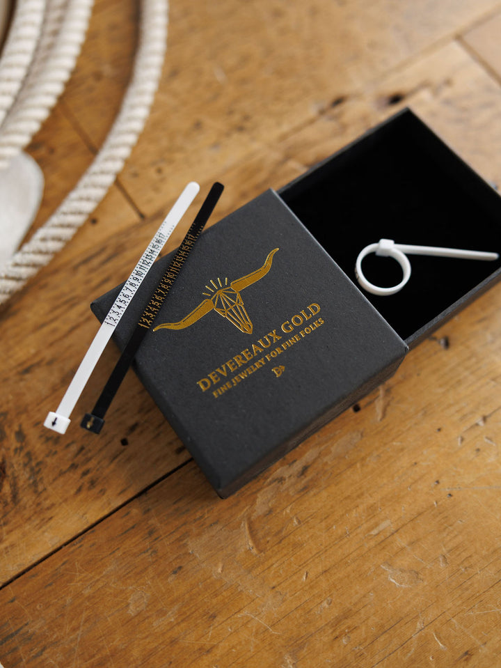 A black jewelry box with a gold Savannah Sevens western life{&}style logo and "Ring Sizing Kit" on the lid sits open on wood, displaying a silver earring, an at-home ring sizing kit, and two monochrome tags atop the box. Collaboration the Devereaux Gold.