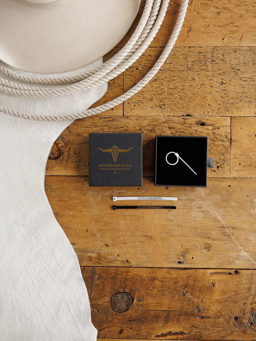 A flat lay on wood displays the Savannah Sevens western life{&}style Ring Sizing Kit: a white cowboy hat, coiled rope, black jewelry box with ring sizer--perfect for at-home ring sizing. Collaboration the Devereaux Gold.