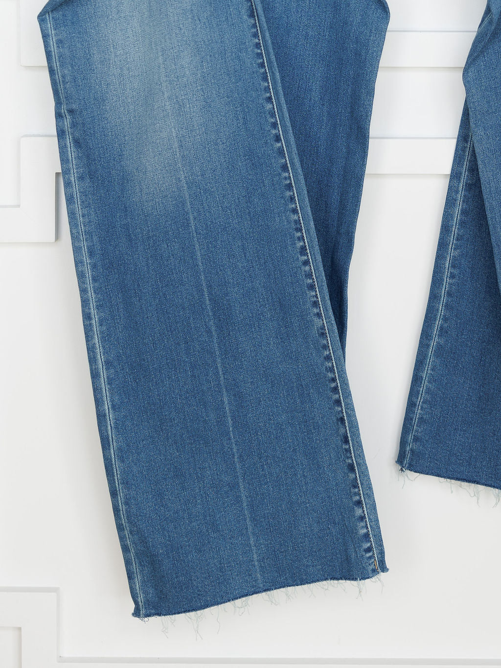 Close-up of the bottom hem of The Jacey High Rise Wide Leg Jean by Savannah Sevens western life{&}style in blue, medium wash denim with a raw, frayed edge, shown against a white background with decorative molding.