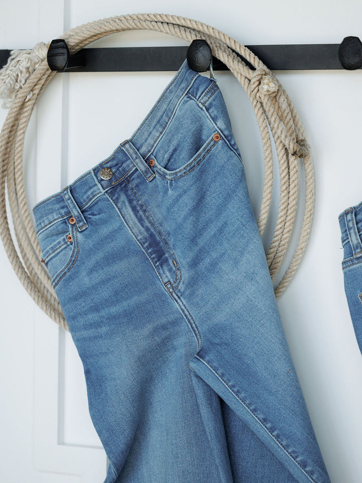 A pair of Savannah Sevens western life{&}style The Jacey High Rise Wide Leg Jeans in light blue hangs on a black wall hook, alongside a coiled rope, set against a white background.