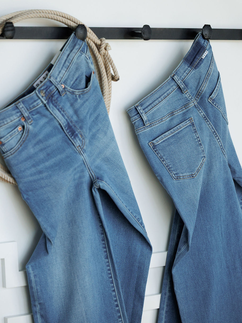 Two pairs of Savannah Sevens western life{&}style jeans hang side by side on black hooks against a white wall, one with a coiled rope. One pair is The Jacey High Rise Wide Leg Jean in medium wash denim.
