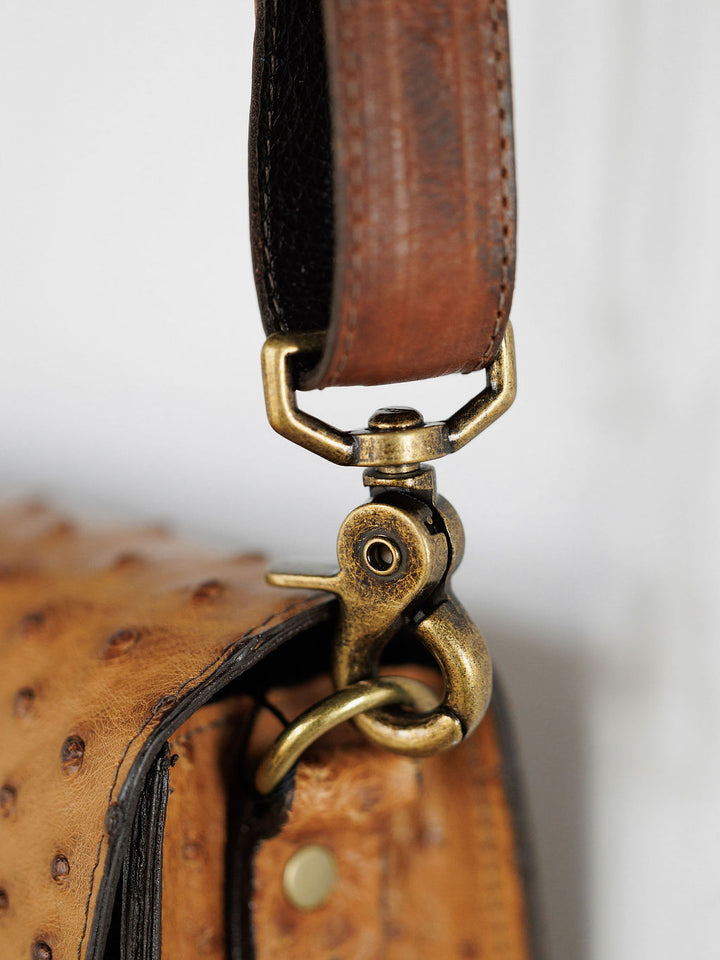A close-up of The Chicota by Savannah Sevens western life{&}style—a saddle tan crossbody bag for women featuring a textured surface and a brass swivel clasp connecting the strap to the bag.