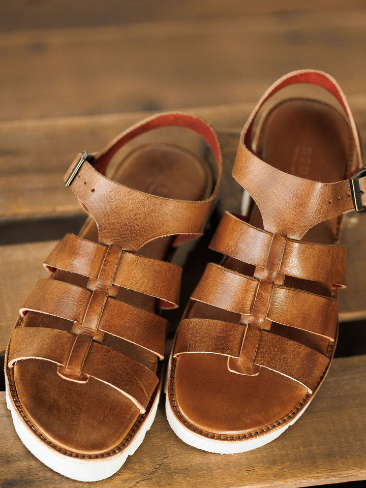 A close-up of The Tuscola brown leather platform sandals by Savannah Sevens western life{&}style, featuring open toes, white soles, multiple straps with a buckle, cushioned insole, and placed on a wooden surface.