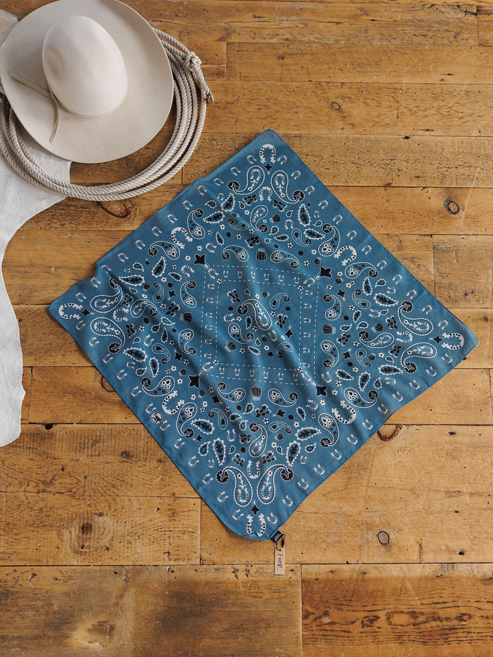 The Lucky Paisley Neck Scarf {Cool Blue} by Savannah Sevens western life{&}style is displayed on a wooden floor beside a beige cowboy hat and coiled rope for a rustic Western-style look.