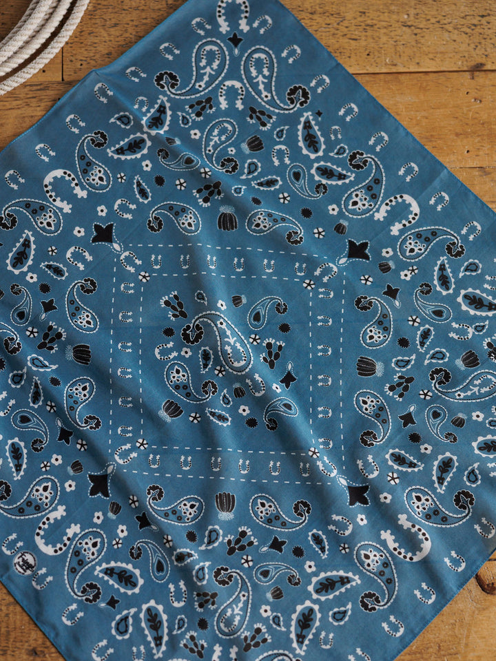 The Lucky Paisley Neck Scarf {Cool Blue} by Savannah Sevens western life{&}style, featuring a white and black paisley print, is displayed flat on wood with a coiled white rope in the upper left corner.