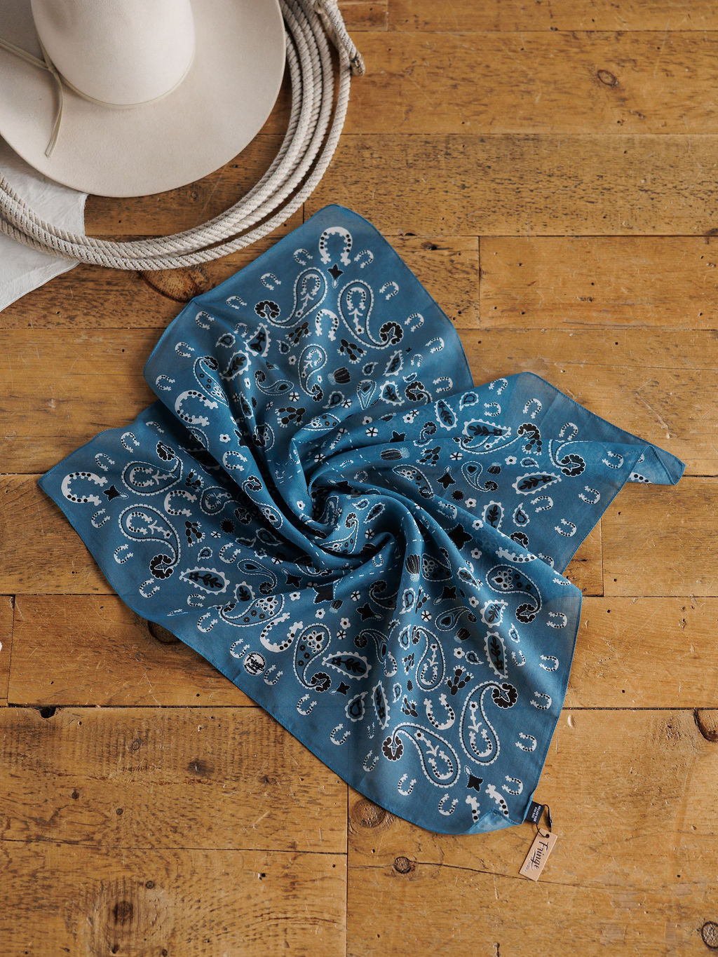 The Lucky Paisley Neck Scarf {Cool Blue} by Savannah Sevens western life{&}style lies on a wooden surface, with a white cowboy hat and coiled rope nearby, adding a western vibe.