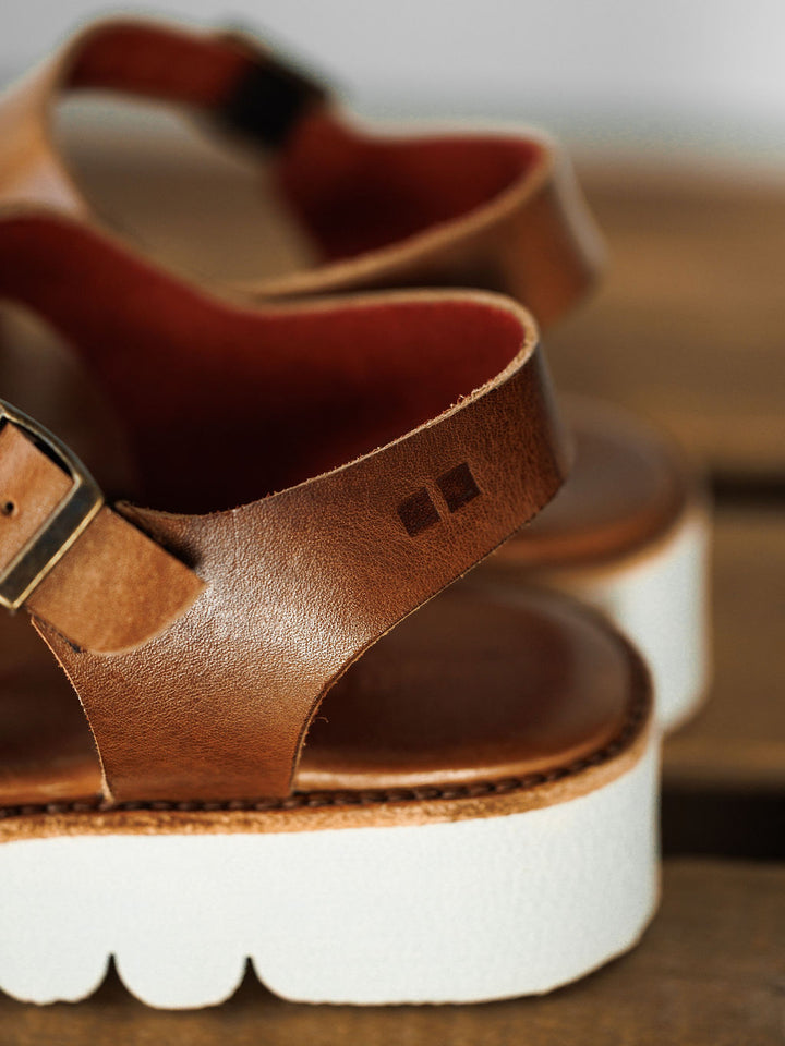Close-up of The Tuscola by Savannah Sevens western life{&}style: a leather platform sandal featuring a white thick sole, cushioned insole, buckle, and small square logo embossed on the strap. Blurred background highlights its texture. Sandals made by BedStu.