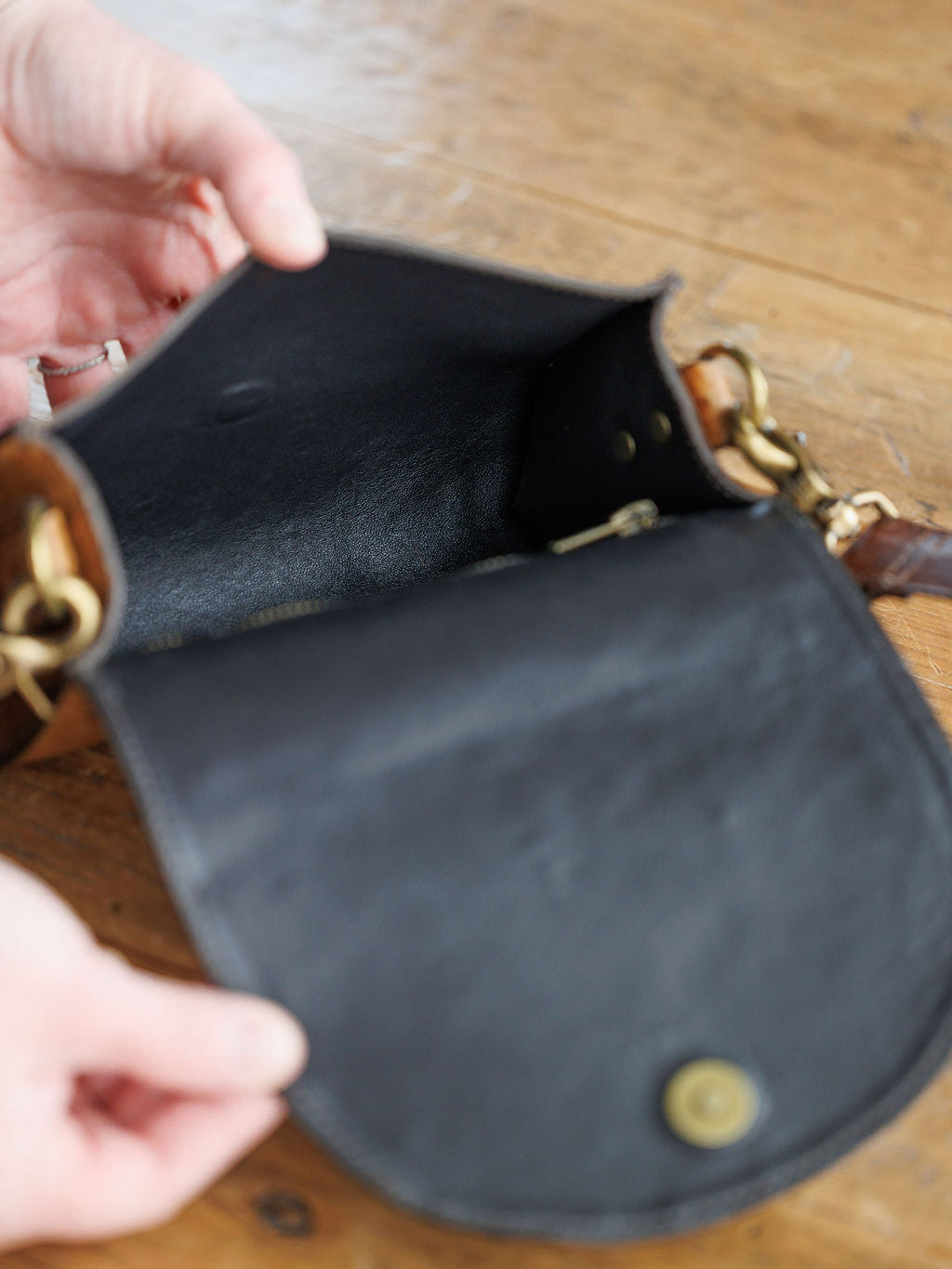 A person holds open the flap of The Chicota by Savannah Sevens western life{&}style, a small black women’s crossbody bag with gold hardware, showing its empty interior on a wooden surface.
