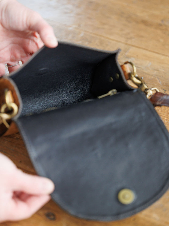 A person holds open the flap of The Chicota by Savannah Sevens western life{&}style, a small black women’s crossbody bag with gold hardware, showing its empty interior on a wooden surface.