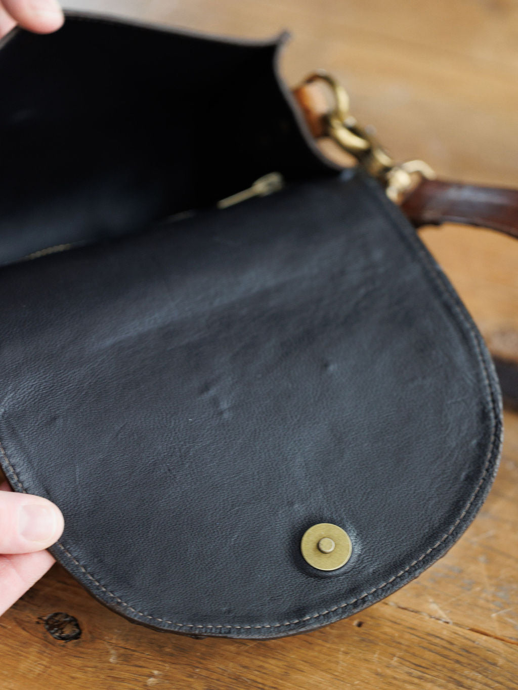 A hand opens The Chicota by Savannah Sevens western life{&}style—a black ostrich leather clutch with a brass snap button. Its interior and part of a saddle tan strap with a brass clasp are visible on a wooden surface.