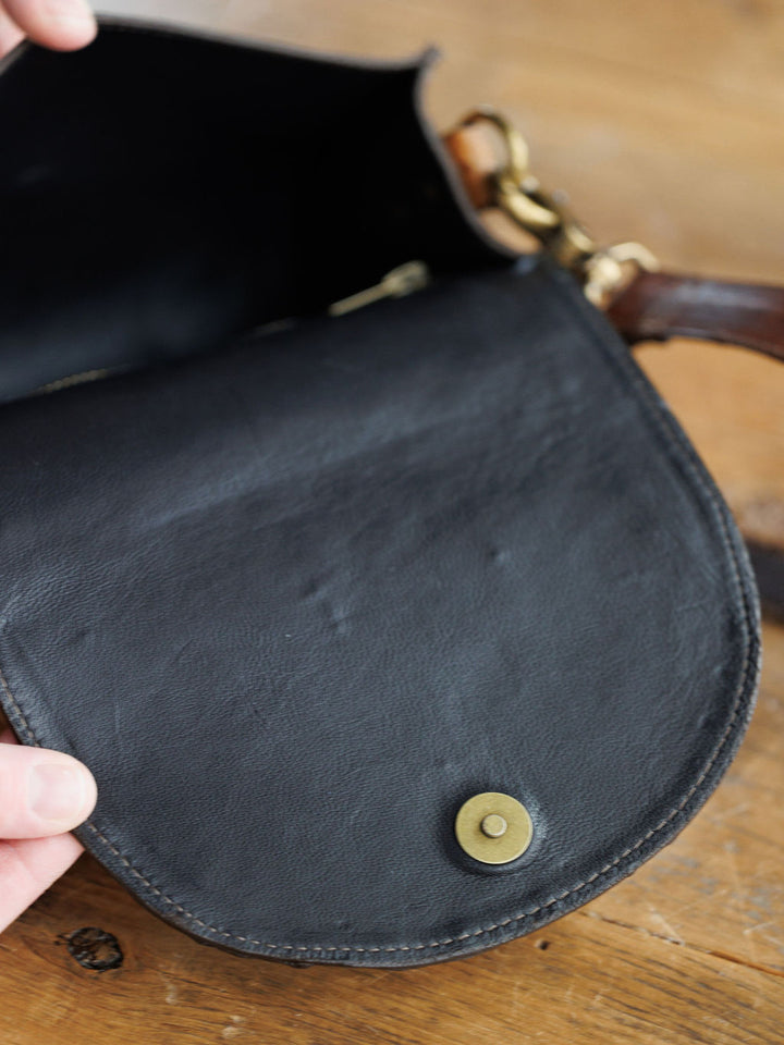 A hand opens The Chicota by Savannah Sevens western life{&}style—a black ostrich leather clutch with a brass snap button. Its interior and part of a saddle tan strap with a brass clasp are visible on a wooden surface.
