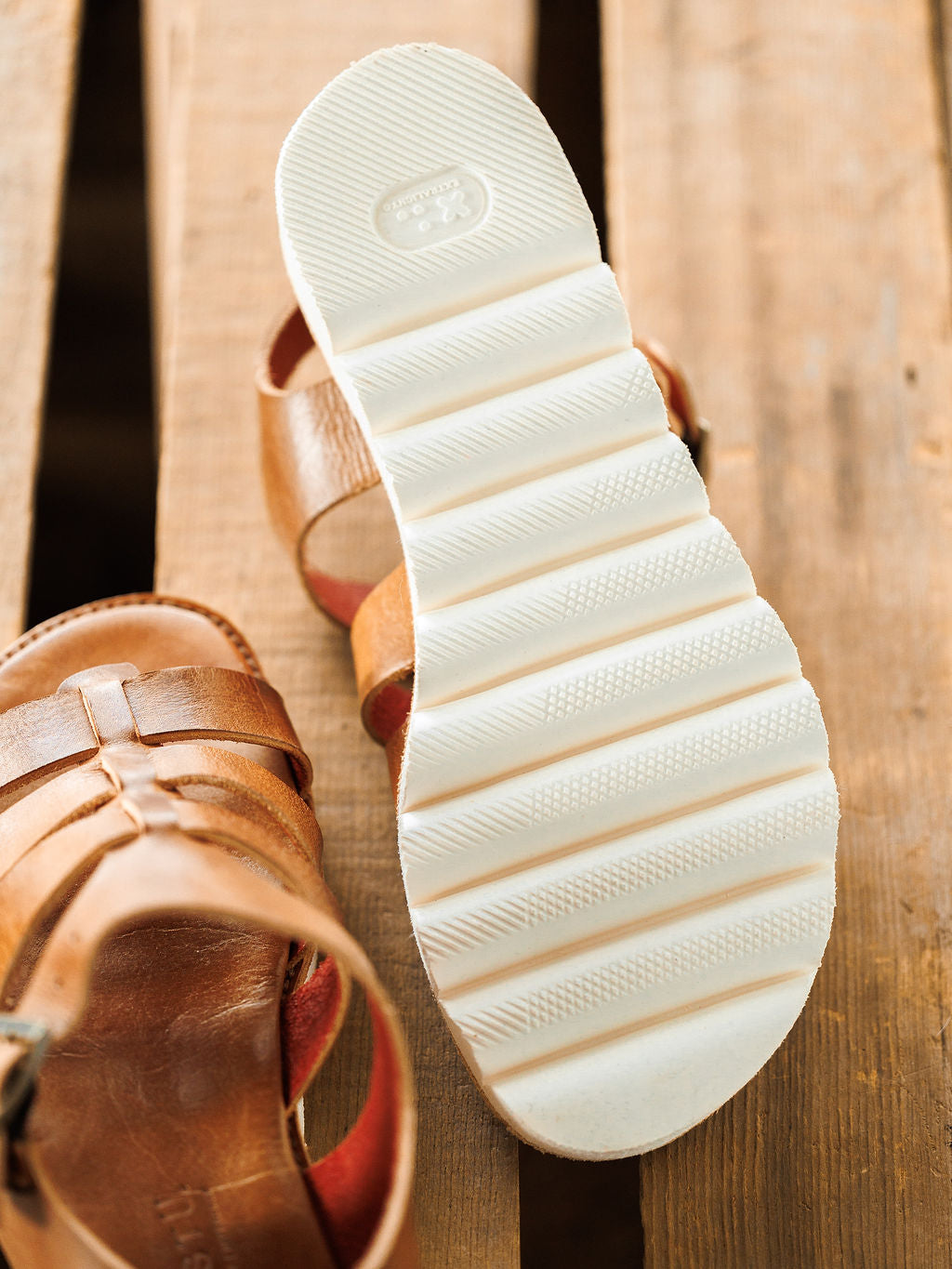 A close-up of Savannah Sevens western life{&}style The Tuscola brown leather platform sandals on wood, with one flipped to show its white, ribbed sole and cushioned insole. Sandals made by BedStu.