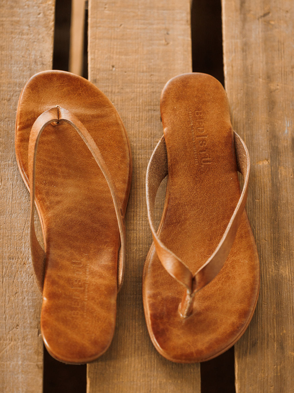 A pair of The Sadler brown leather flip flop sandals by Savannah Sevens western life{&}style, featuring a cushioned footbed, are placed side by side on a wooden surface. Sandals made by BedStu. 