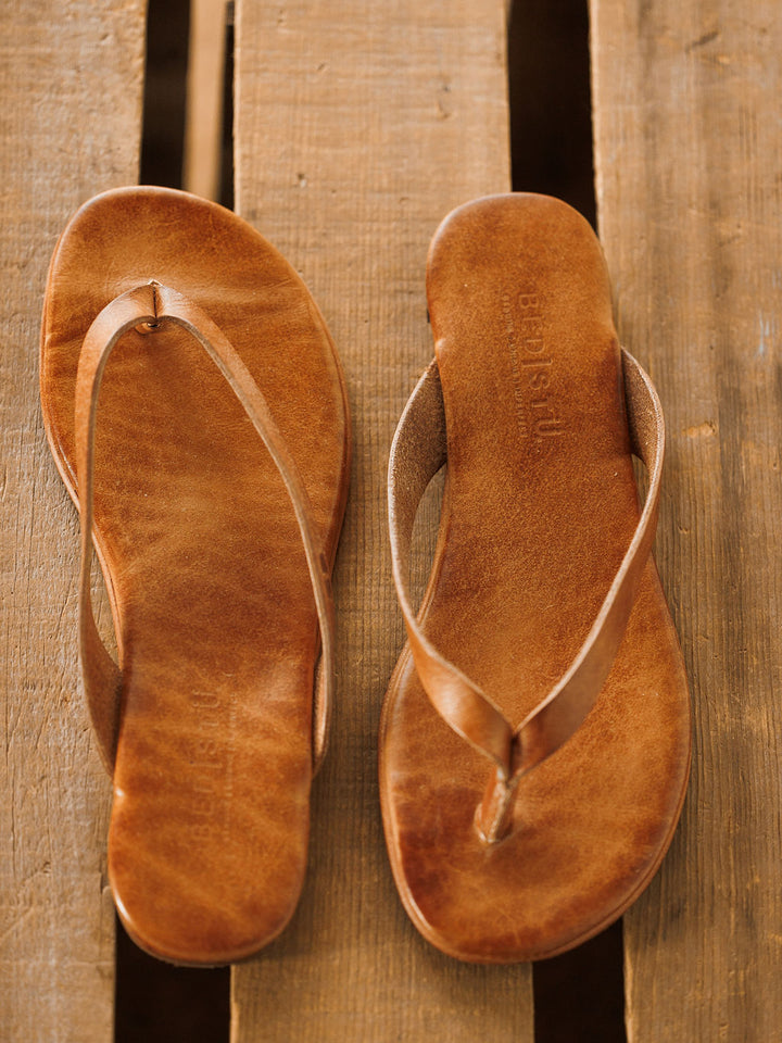 A pair of The Sadler brown leather flip flop sandals by Savannah Sevens western life{&}style, featuring a cushioned footbed, are placed side by side on a wooden surface. Sandals made by BedStu. 