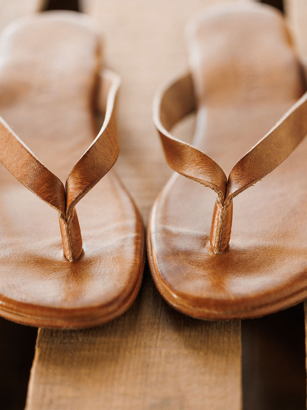 Close-up of The Sadler brown leather flip flops by Savannah Sevens western life{&}style, featuring toe straps and a cushioned footbed, resting on a wooden surface.