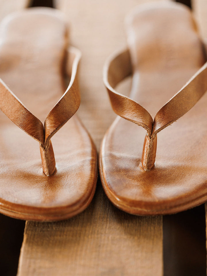 Close-up of The Sadler brown leather flip flops by Savannah Sevens western life{&}style, featuring toe straps and a cushioned footbed, resting on a wooden surface.