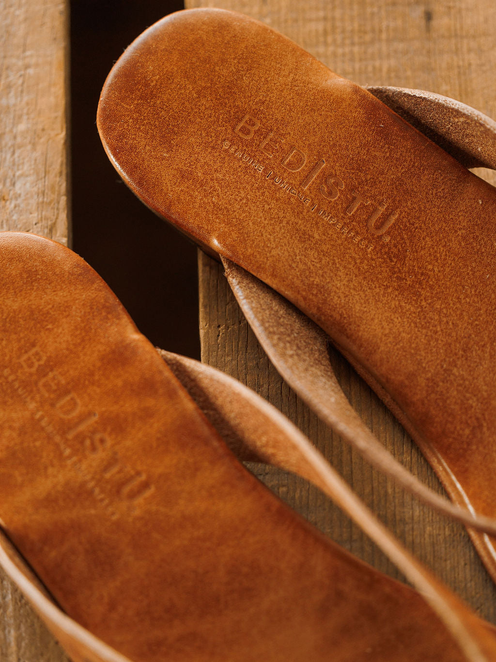 Close-up of two brown The Sadler flip flops by Savannah Sevens western life{&}style on a wooden surface, with the brand embossed on the insole. The texture, craftsmanship, and cushioned footbed are clearly visible. Sandals by BedStu.