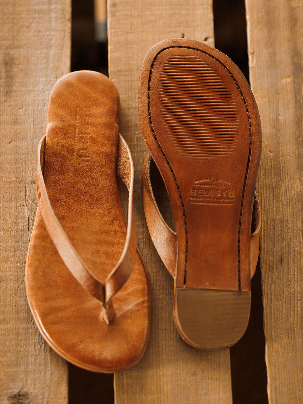 A pair of The Sadler tan leather flip flop sandals by Savannah Sevens western life{&}style, featuring embossed logos on the cushioned footbed and sole, displayed on a rustic wooden surface. Sandals by BedStu.