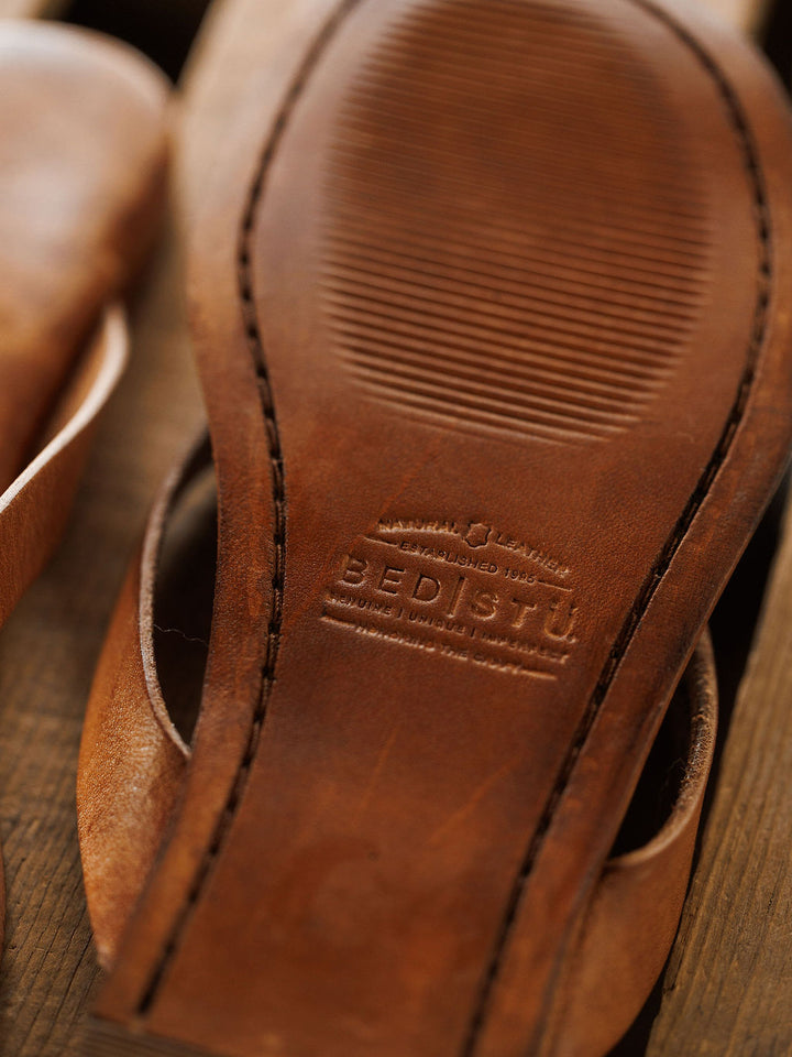 Close-up of The Sadler by Savannah Sevens western life{&}style: a brown leather shoe sole with visible stitching, embossed "Genuine Leather" text, and a textured pattern suggesting quality and a cushioned footbed for comfort. Sandals by BedStu.