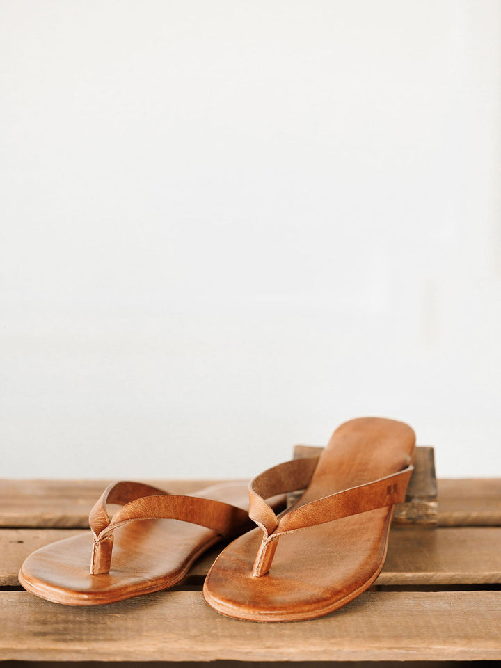 A pair of The Sadler leather flip flops by Savannah Sevens western life{&}style, featuring a cushioned footbed, rests on a wooden surface against a plain, light background. Sandals by BedStu.