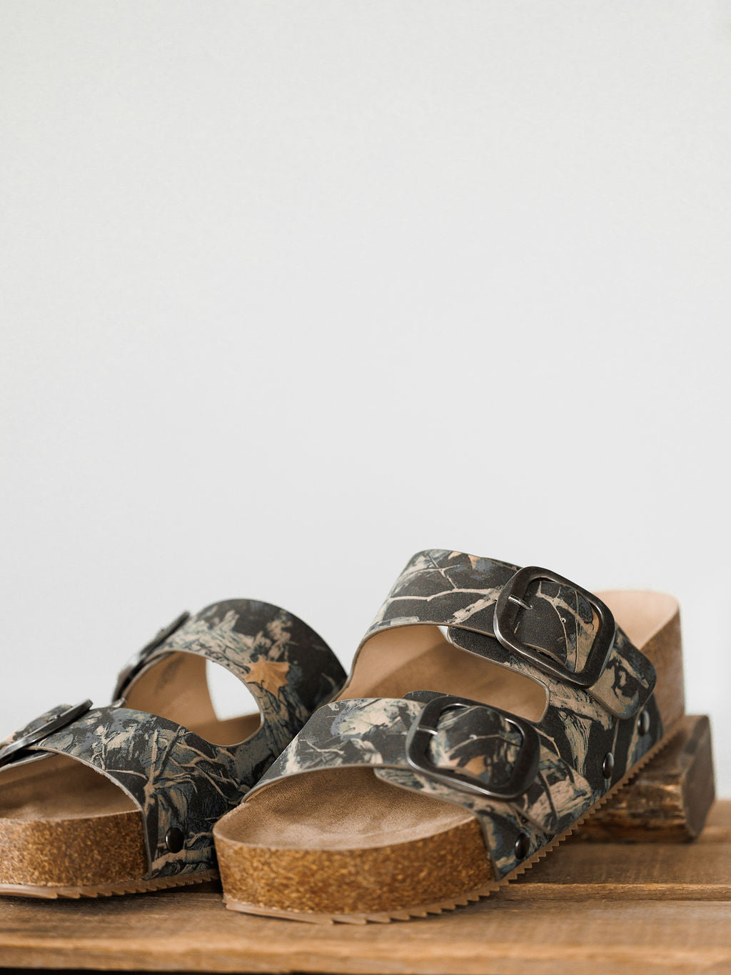 The Kane by Savannah Sevens western life{&}style features camo slip-on sandals with cork soles, cushioned insoles, and two wide camouflage straps with large black buckles, shown on a wooden surface against a plain background.