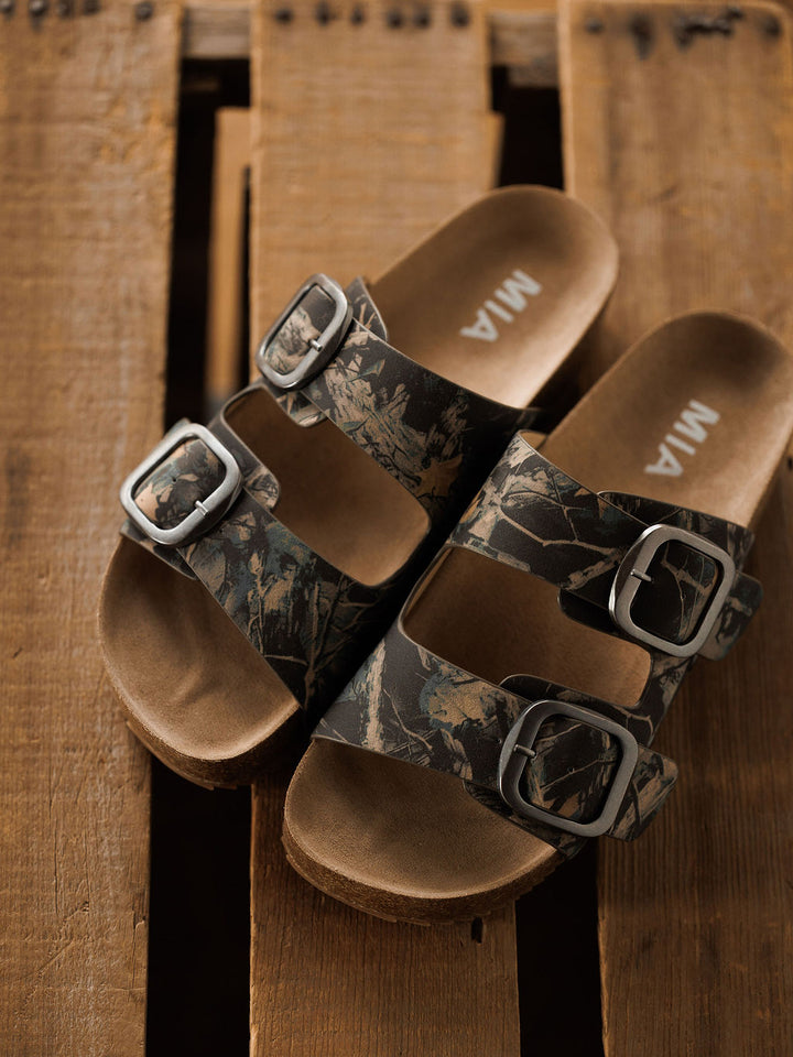 A pair of The Kane slip-on sandals by Savannah Sevens western life{&}style with brown cork soles, camo-patterned straps, silver buckles, and cushioned insoles rests on a rustic wooden surface.