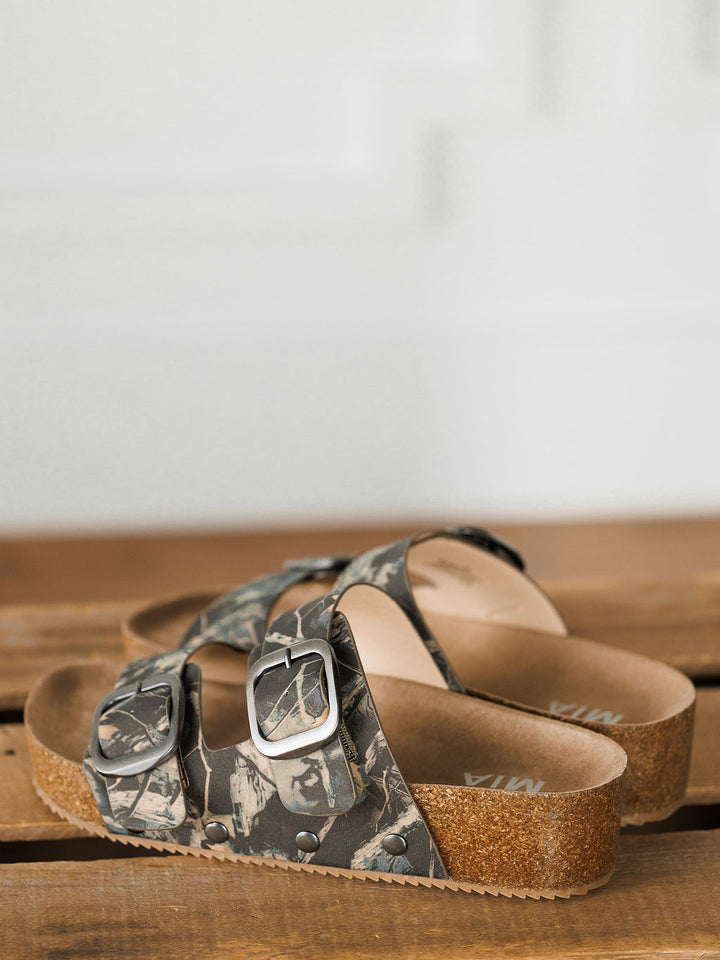 The Kane by Savannah Sevens western life{&}style: camo slip-on sandals with cork soles, silver buckles, and cushioned insoles for comfort, displayed on a wooden surface.