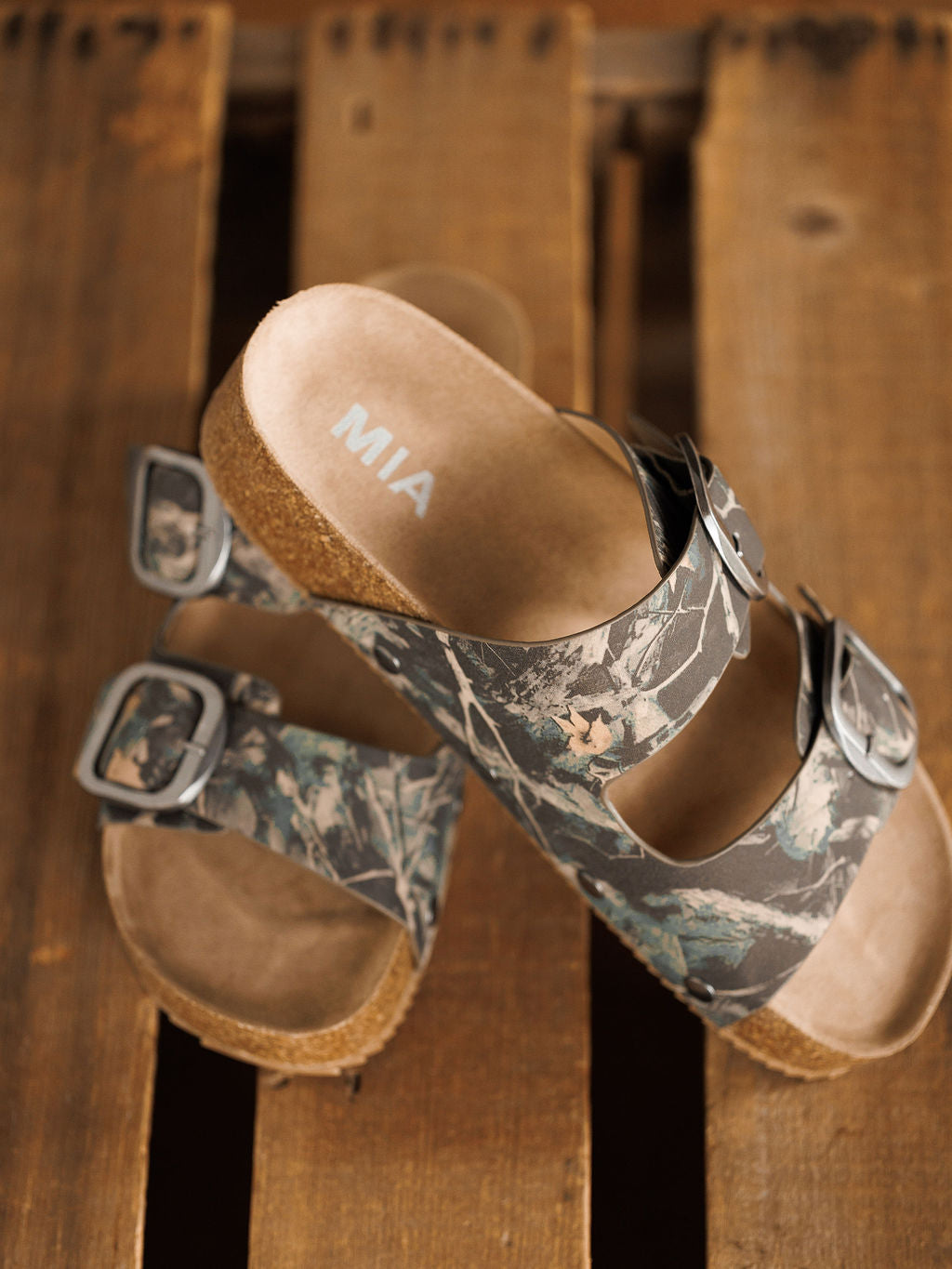A pair of camo slip-on sandals called The Kane by Savannah Sevens western life{&}style, featuring cork soles, cushioned insoles, and silver-buckled camouflage straps, displayed on a rustic wooden surface. Brand name printed on footbed.
